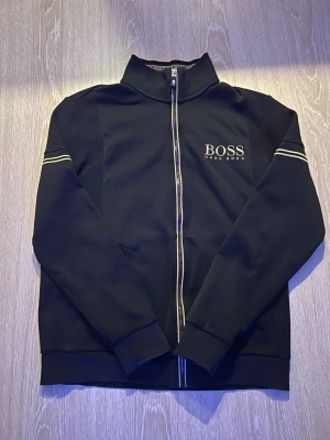 Svart ziptröja från Hugo Boss - Snygg svart ziptröja från Hugo Boss med vit logga på bröstet och vita detaljer längs dragkedjan och ärmarna. Tröjan har hög krage, ribbade muddar och sportig design. Perfekt för en stilren och avslappnad look. Storlek M men passar som XS 