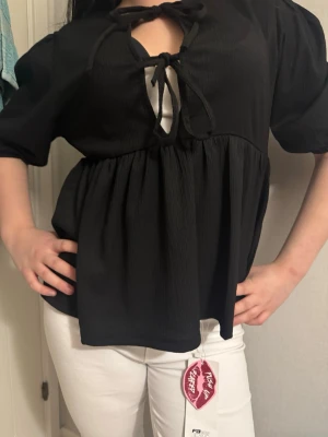 Svart blus med knyt och puffärm - Trendig svart blus med korta puffärmar och två knytband framtill som skapar en snygg cut-out detalj vid halsen. Blusen har en lös, flowy passform och är tillverkad i ett mjukt, strukturerat tyg. Perfekt att styla med jeans eller kjol för en cool look.