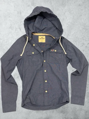 Grå skjorta med huva från Hollister - Snygg grå skjorta från Hollister med huva och vita dragsnören. Skjortan har långa ärmar, två bröstfickor med knappar och guldfärgade knappar framtill. Tillverkad i mjuk bomull, perfekt för en avslappnad streetstil. Mått: Bredd; 40 cm. Längd; 62 cm. Ärm; 72 cm.