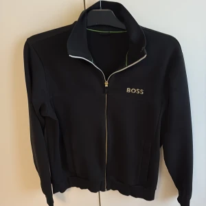 Svart ziptröja från BOSS - Svart långärmad ziptröja från BOSS med guldfärgad dragkedja och broderad logga på bröstet. Tröjan har hög krage och ribbade muddar. Perfekt för en stilren och sportig look.