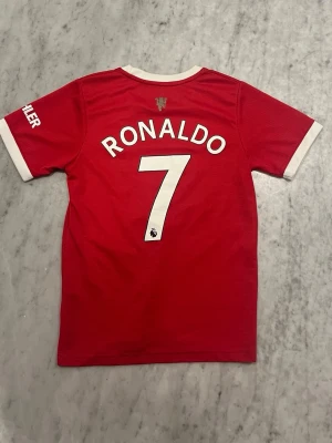 Manchester United Ronaldo matchtröja - Röd Manchester United matchtröja med vita detaljer, tryck med Ronaldo och nummer 7 på ryggen. Premier League-logga på siffran och klubbens emblem i nacken. Tillverkad i lätt och ventilerande material, perfekt för fotbollsträning eller match.