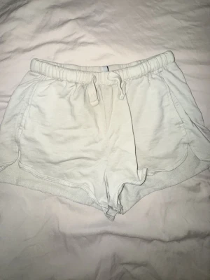 beige mjukisshorts  - Säljer ett par vita mjukisshorts från H&M Basic med snörning i midjan och två sidofickor. Shortsen är tillverkade i en mjuk bomulls- och polyestermix och har en avslappnad passform. Perfekta för sommardagar eller chill hemma.