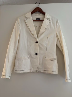 Vit kavaj från Maria Westerlind - Vit kavaj från Maria Westerlind med svarta sömmar och två knappar framtill. Klassisk krage och två fickor fram. Materialet är bomull. Perfekt för en trendig och clean look passar till allt möjligt även utklädnad 