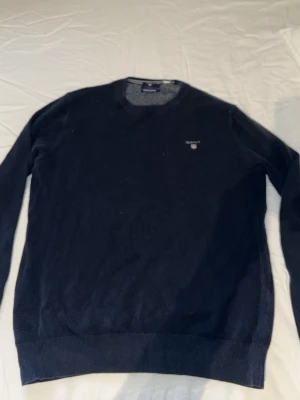 Mörkblå stickad tröja från Gant - Snygg mörkblå stickad tröja från Gant i premium bomull. Tröjan har rund halsringning, diskret Gant-logga broderad på bröstet och ribbade muddar vid ärmslut och nederkant. Perfekt för en stilren och klassisk look.