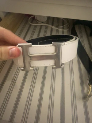 Vitt läderbälte med H-spänne - Snyggt vitt bälte i läder med ett stort, silverfärgat H-format spänne. Bältet är svart på insidan och har stilrena sömmar längs kanterna. Perfekt accessoar för att lyfta vilken outfit som helst. 110cm