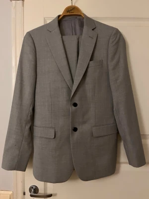Ljusgrå kostym från Dressmann - Snygg ljusgrå kostym från Dressmann i slim fit-modell. Klassisk kavaj med två knappar, två fickor med lock och bröstficka. Tillverkad i ett mjukt material som ger en stilren look. Perfekt för dig som vill ha en modern och elegant stil. Endast använd en gång.