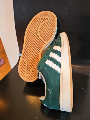 Gröna Adidas sneakers i mocka - Snygga gröna Adidas sneakers i mocka med klassiska vita ränder på sidorna och beige sula. Låg modell med retrovibbar och tydlig yttersula i gummi. Perfekta för dig som gillar streetstyle och vill ha något som sticker ut. De är ljusare men är lite blötta på bilden