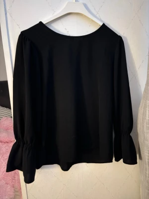 Svart blus med vida ärmar - Stilren svart blus med rund halsringning och långa, vida ärmar som har rynkade muddar. Blusen har en knapp i nacken för enkel påklädning. Perfekt till jeans eller kjol för en trendig look.