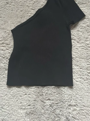 Svart oneshoulder topp - Basic svart oneshoulder topp med ribbad struktur. Toppen har en kort ärm på ena sidan och är ärmlös på andra, vilket ger en unik och trendig look. Säljer då den är för liten, använd men bra skick :))