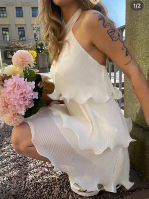 Missäy sommar klänning - Superfin vit kortklänning med halterneck och flera lager volanger. Klänningen är ärmlös och har en flowy, luftig känsla som passar perfekt till varma dagar. Materialet är lätt plisserat och ger en snygg struktur.