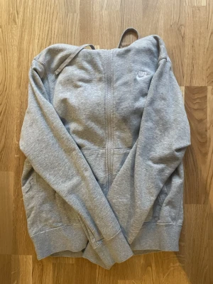 Grå zip hoodie från Nike - Snygg grå zip hoodie från Nike med vit logga på bröstet. Hoodien har dragkedja framtill, två stora fickor och justerbar huva med snören. Tillverkad i mjukt sweatshirtmaterial som är perfekt för chill eller streetwear.