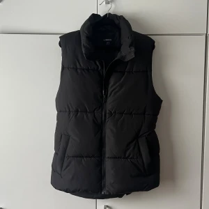 Svart dunväst från Lindex - Snygg svart dunväst från Lindex med hög krage och dragkedja framtill. Västen har två fickor på sidorna och ett bälte i midjan för en mer markerad siluett. Perfekt för lager på lager och enkel att styla till olika outfits.