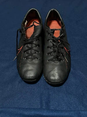 Svarta Nike fotbollsskor med röd detalj - Säljer ett par svarta Nike fotbollsskor med röda detaljer och klassisk Nike-swoosh på sidan. Skorna har snörning och en låg profil, samt röd insida. Materialet är syntetiskt och designen är slimmad för bra bollkontroll på planen.