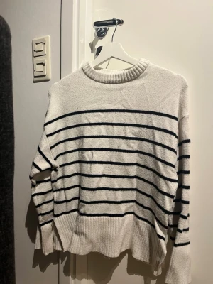 Vit stickad tröja med svarta ränder - Säljer en vit stickad tröja med svarta horisontella ränder. Tröjan har en bred ribbad mudd vid hals, ärmslut och nederkant samt en avslappnad passform. Perfekt till jeans för en enkel och snygg look.