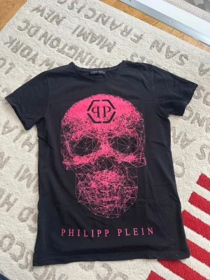 Svart Philipp Plein t-shirt med rosa tryck - Cool svart t-shirt från Philipp Plein med ett stort grafiskt rosa dödskalle-tryck på framsidan och logga ovanför. T-shirten har rund hals och korta ärmar. Perfekt för dig som gillar streetwear och statement-plagg.