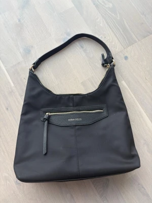 Svart handväska från Anna Field - Cool svart handväska från Anna Field med guldfärgade dragkedjor och en praktisk ytterficka. Väskan har en enkel, slät design och är gjord i ett tåligt syntetmaterial. Perfekt axelrem som gör den enkel att bära. Rymmer allt du behöver för dagen.