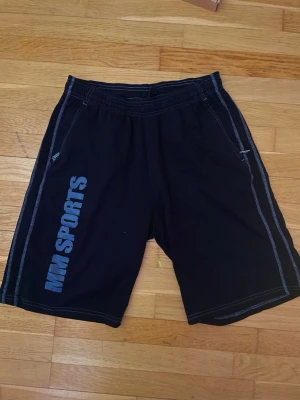 MM Sports Svarta träningsshorts - Svarta träningsshorts från MM Sports med vit kontrastsöm och tryckt logga längs benet. Shortsen har elastisk midja och två sidofickor. Perfekta för gymmet eller annan träning.