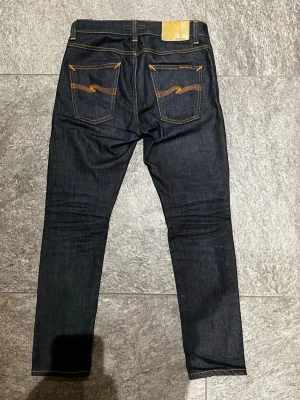 Mörkblå jeans från Nudie Jeans - Säljer ett par mörkblå jeans från Nudie Jeans med klassisk femficksmodell och kontrastsömmar. Jeansen har smal passform och detaljerade bakfickor med Nudie Jeans signaturbrodyr. Tillverkade i rå denim för en stilren look. (Jeansen är inte missfärgade, om det ser mörkare ut så är det skuggor)