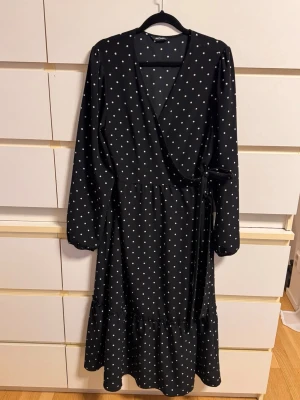Svart prickig omlottklänning - Snygg svart omlottklänning från MONKI med vita prickar och knyt i midjan. Klänningen har lång ärm, v-ringad hals och volang nedtill. Perfekt för dig som gillar en feminin och trendig look.