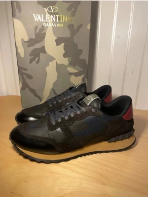 Valentino Garavani camo sneakers - Snygga sneakers från Valentino Garavani med camomönster i svart, blått och grått. Skorna har svarta mockadetaljer, mörkblå partier och en röd detalj på hälen. Yttermaterialet är en mix av läder och textil, med robust sula och grå skosnören.