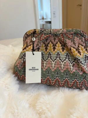 Mönstrad kuvertväska från Beck Söndergaard - Säljer en unik kuvertväska från Beck Söndergaard med färgstarkt mönster i rosa, gult, grönt och svart. Väskan har en mjuk, tygklädd yta och klassisk form med lätt puffig look. Perfekt accessoar för att lyfta din outfit med lite extra färg och stil.