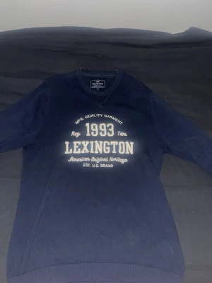 Mörkblå Lexington tröja med tryck - Mörkblå tröja från Lexington med vitt tryck framtill där det står '1993 Lexington American Original Heritage'. Tröjan har V-ringning och långa ärmar. Perfekt för dig som gillar klassisk college-stil.