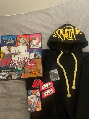 Svart hoodie från Syna World - Svart hoodie från Syna World med stor gul logga i graffiti-stil på huvan och tjocka, tvinnade gula snören. Hoodien har klassisk passform och en stor huva. Perfekt för dig som gillar streetwear och vill sticka ut med coola detaljer.