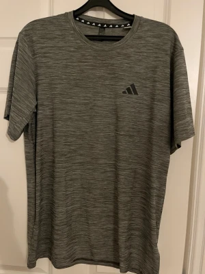 Gråmelerad t-shirt från Adidas - Snygg gråmelerad t-shirt från Adidas med diskret logga på bröstet. T-shirten har rund halsringning och korta ärmar. Tillverkad i ett mjukt och lätt material som passar perfekt till vardags eller träning.