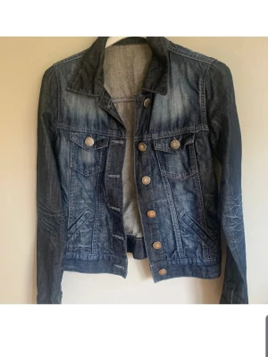 Mörkblå klassisk jeansjacka - Snygg mörkblå jeansjacka med slitningar och kopparfärgade knappar. Två bröstfickor med lock och knappar samt två sidofickor. Jackan har en klassisk krage och figurnära passform, perfekt till alla dina outfits.