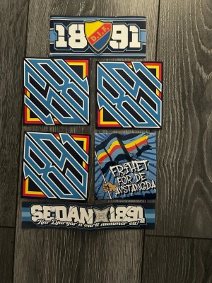 Djurgården klistermärken paket - Paket med flera klistermärken från Djurgårdens IF. Motiven inkluderar DIF-skölden, årtalet 1891, grafiska mönster i blått, gult och rött samt texten 'Frihet för de avstängda'. Perfekt för att visa ditt stöd för laget.