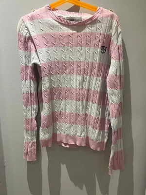 Rosa och vit kabelstickad tröja Gant - Säljer en kabelstickad tröja från Gant med breda rosa och vita ränder. Tröjan har rund hals, långa ärmar och ett broderat Gant-emblem på bröstet. Materialet är mjukt och bekvämt, perfekt för att hålla sig varm. Passar dig som gillar klassisk preppy-stil.