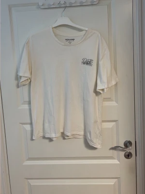 Vit t-shirt från Jack & Jones - Säljer en vit t-shirt från Jack & Jones med trycket 'Dark Manifesto' och en stor grafisk illustration på ryggen. T-shirten har rund hals, korta ärmar och är gjord i mjuk bomull. Perfekt för dig som gillar streetwear vibes och coola prints.