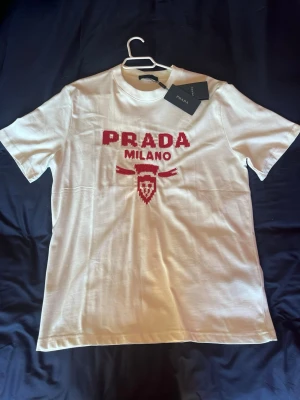 Vit t-shirt från Prada med röd logga - Snygg vit t-shirt från Prada med röd broderad PRADA MILANO-logga och emblem på bröstet. Klassisk passform och rund hals. Tillverkad i mjuk bomull för en skön känsla. Perfekt för dig som vill ha en stilren och exklusiv look. Säljs då den aldrig används. 