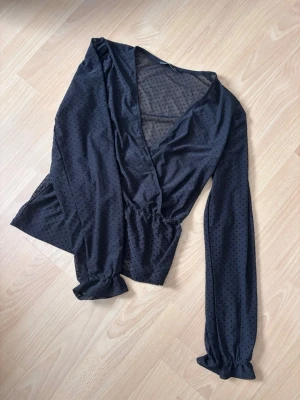 Svart prickig omlottblus - Trendig svart omlottblus med små diskreta prickar och transparent mesh upptill. Blusen har långa ärmar med resår vid ärmslut och en smickrande v-ringning. Perfekt för att lyfta vilken outfit som helst.