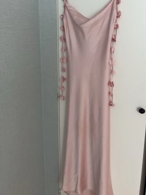 Ljusrosa långklänning med blom-snören  - Säljer en ljusrosa långklänning i satinliknande material med tunna axelband och blom-band längst sidorna, använd en gång 💞💞storlek 34/xs
