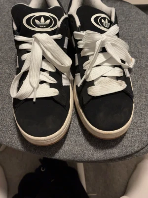 Svarta Adidas Campus sneakers - Klassiska Adidas Campus sneakers i svart mocka med vita detaljer och vita snören. Skorna har den ikoniska tre ränder-designen på sidan och Adidas-logga på plösen. Låg modell med vit sula och bekväm passform. Perfekta för en streetwear-look. Använda 1 gång 🥰