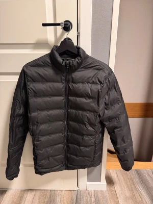 Svart pufferjacka med dragkedja - Snygg svart pufferjacka med ståkrage och horisontella sömmar. Jackan har hel dragkedja framtill och är lätt vadderad för extra värme. Perfekt för kalla dagar och enkel att matcha med olika outfits.