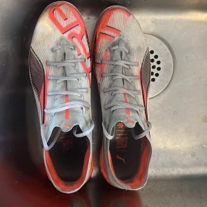 Puma grå-röda fotbollsskor - Säljer ett par gråa fotbollsskor från Puma med röda och svarta detaljer. Skorna har snörning, låg profil och en sportig design med Pumas logga på sidan. Insidan är röd och ovandelen är tillverkad i ett syntetmaterial som ger bra bollkontroll.