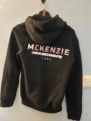 Svart hoodie från McKenzie - Snygg svart hoodie från McKenzie med vit och röd logga på bröstet och stor text på ryggen. Klassisk känguruficka framtill, justerbar huva med snören och ribbade muddar. Perfekt för en avslappnad och sportig stil. Först till kvarn som gäller passar dig som har storlek XS. Säljer den eftersom den inte passar mig längre