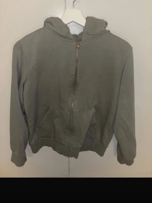 Grå zip hoodie med fickor - En enkel grå hoodie med dragkedja framtill och kängurufickor. Tröjan har huva med dragsko och ribbade muddar vid ärmar och nederkant. Perfekt för en avslappnad stil och funkar året runt. Materialet känns mjukt och bekvämt.