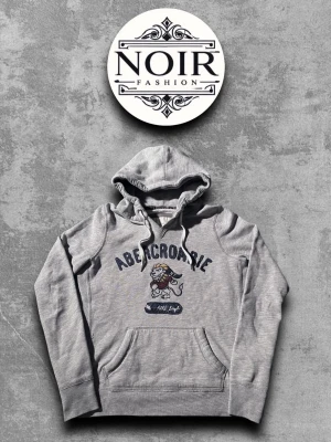 Grå hoodie från Abercrombie - Snygg grå hoodie från Abercrombie med tryck av en lejonmaskot och texten 'Abercrombie' på bröstet. Klassisk känguruficka framtill och justerbar huva med dragsko. Mjuk bomullsblandning som känns skön mot huden. Perfekt för en avslappnad streetwear-look.