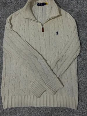 Vit kabelstickad tröja Polo Ralph Lauren - Kabelstickad vit tröja från Polo Ralph Lauren med halv dragkedja och klassisk blå logga broderad på bröstet. Tröjan har ribbade muddar och krage, samt är gjord i mjukt material som känns lyxigt och bekvämt. Perfekt för dig som gillar stilrena och tidlösa plagg.
