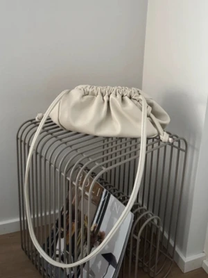 Krämvit axelremsväska med dragsko - Säljer en krämvit handväska i mjukt konstläder med dragskostängning och lång axelrem. Väskan har en puffig, modern form och minimalistisk design som passar perfekt till trendiga outfits. Enkel att matcha och superpraktisk för vardagens essentials.