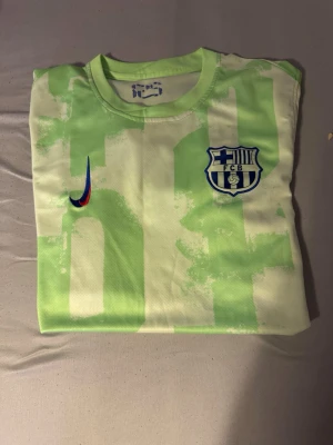 Grön Barcelona fotbollströja Nike - Snygg Barcelona fotbollströja i ljusgrönt med vita inslag och klubbmärke på bröstet. Tröjan har rund hals och är tillverkad i ett lätt, ventilerande material som passar perfekt till träning eller match. Nike-logga i regnbågsfärg på framsidan.