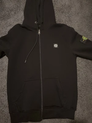 Svart hoodie från Stone Island - Säljer en svart hoodie från Stone Island med dragkedja, huva med dragsko och klassisk logopatch på vänster ärm. Hoodien har två stora fickor framtill och är gjord i mjukt bomullsmaterial. Perfekt för dig som gillar streetwear och snygga detaljer. storlek L men kan passa även Storlek M pris kan diskuteras vid snabb affär