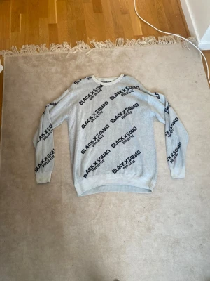 Grå tröja från Black Squad XL - Säljer en ljusgrå tröja från Black Squad i regular fit, storlek XL. Tröjan har svart textmönster med 'BLACK SQUAD' tryckt över hela plagget och ribbade muddar vid hals, ärmslut och nederkant. Perfekt för dig som gillar streetstyle.
