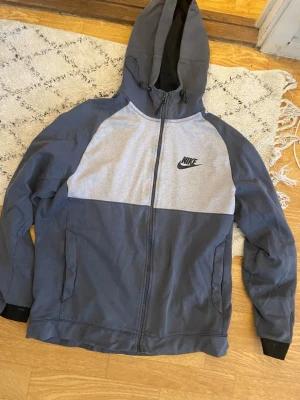 Grå och mörkgrå Nike hoodie - Snygg hoodie från Nike i två nyanser av grått med dragkedja framtill och svart Nike-logga på bröstet. Hoodien har huva med dragsko och muddar vid ärmsluten. Perfekt för en avslappnad och sportig stil.