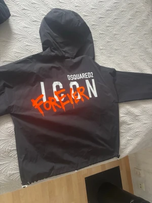 Svart vindjacka Dsquared2 ICON - Svart LIMITED EDITON vindjacka från Dsquared2 med huva och tryck på ryggen. Stor vit text 'ICON' och 'FOREVER' i orange graffiti-stil samt Dsquared2-logga. Lätt material, perfekt för streetwear och trendig look. Äkta samt helt nytt skick