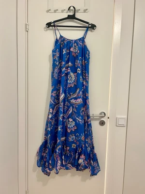 Blommig blå bomull långklänning från MNG - Säljer en blå långklänning från MNG med färgglatt blommönster i rosa, beige och lila. Klänningen har tunna axelband, volangkant nertill och en luftig passform. Perfekt för varma dagar och somriga tillfällen. 100% bomull 