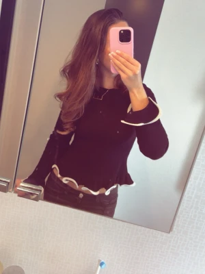 Svart långärmad topp från Ginatricot - Svart långärmad topp från Ginatricot med vita volangdetaljer vid ärmslut och nederkant. Toppen har en figurnära passform och är tillverkad i ett stretchigt, ribbat material som ger en snygg siluett. Perfekt att styla med både jeans och kjol. Den är använd cirka 3 gånger och är i super fint skick och inga skador eller tecken på användning. Slutsåld och köptes för 449 kr💗💗